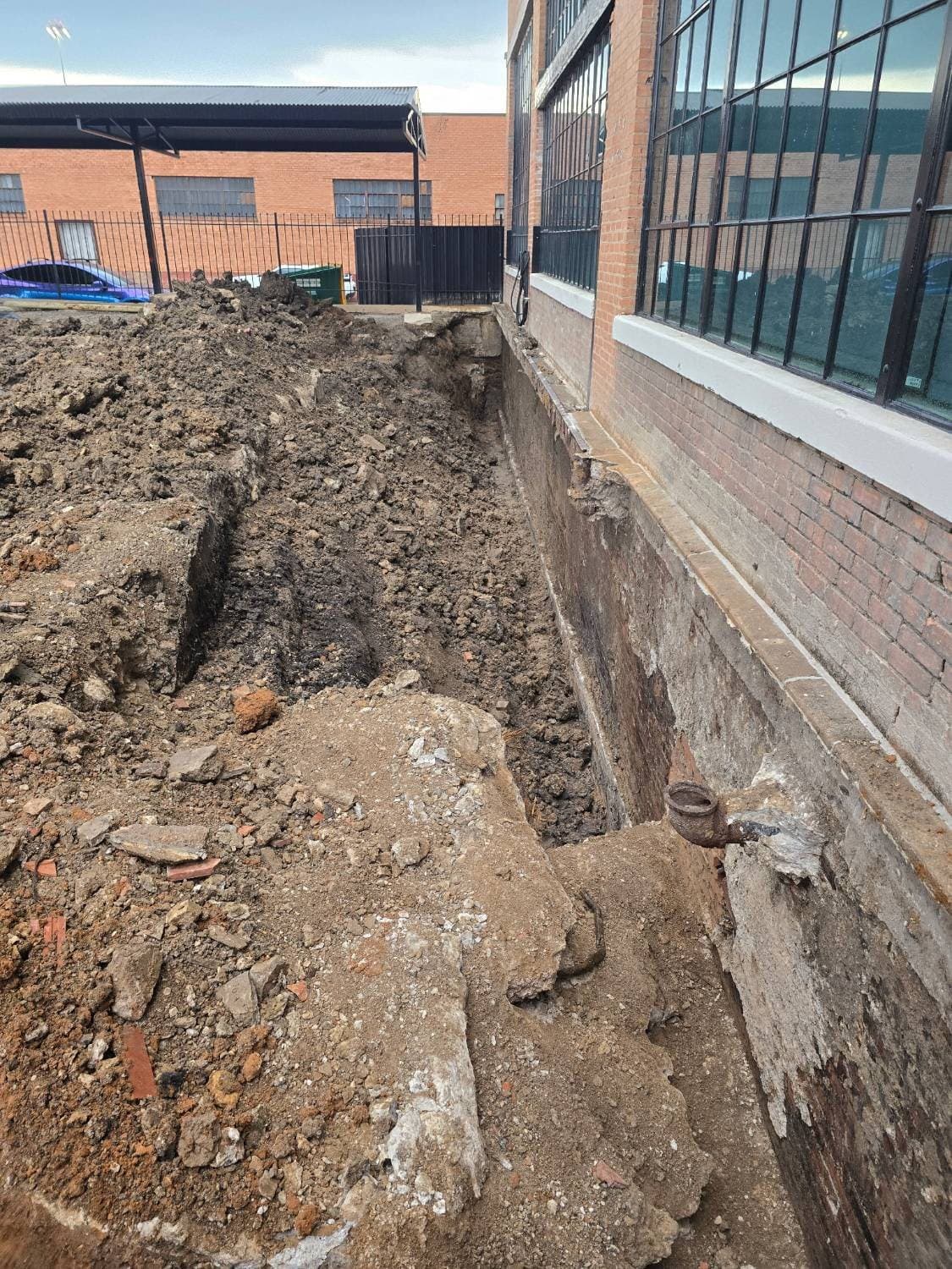 Excavated trench along the brick foundation wall of a converted industrial building, exposing the wall for below-grade waterproofing