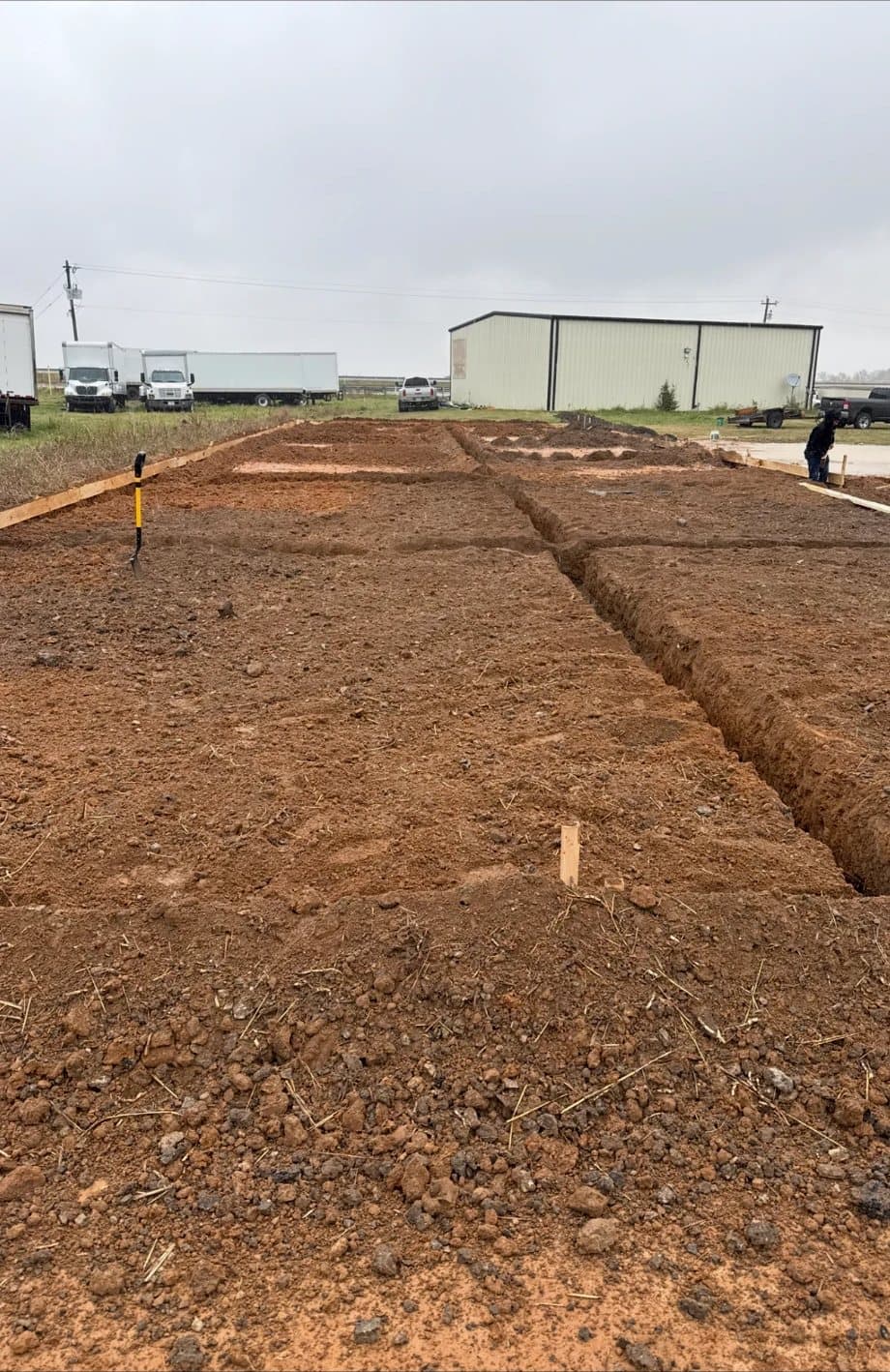 Graded red-clay site with forms and footing trenches laid out next to an existing metal building
