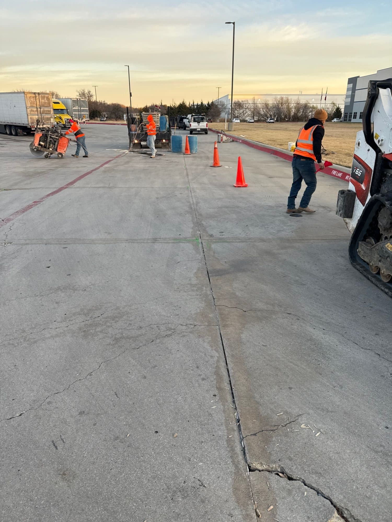 Crew saw-cutting and sealing joints across a concrete truck lot beside a distribution warehouse, with a compact track loader in the foreground