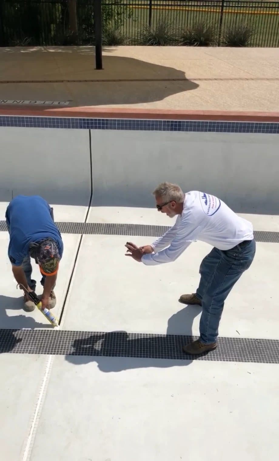 Two workers inside a drained commercial pool, one applying sealant to a floor joint between tile bands