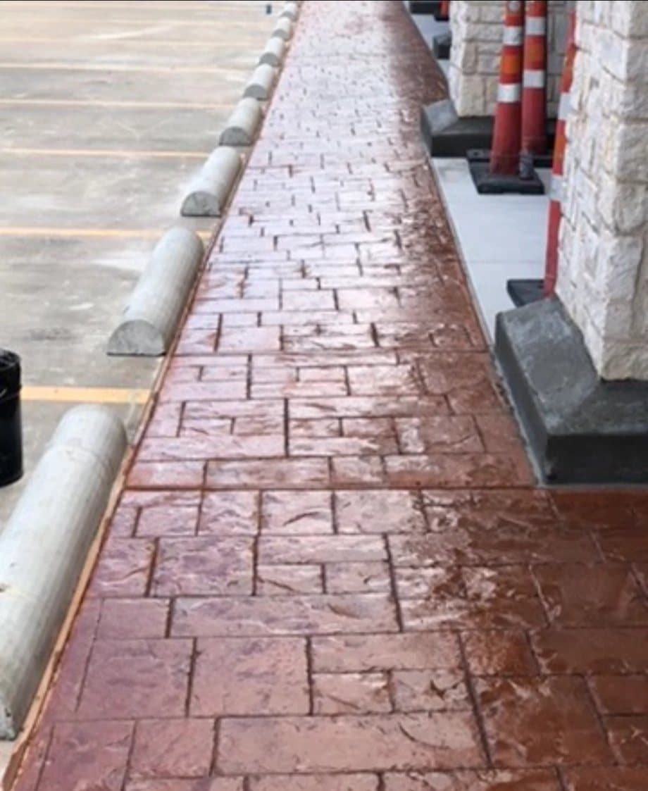 Finished red stamped-concrete walkway along a retail storefront with stone columns and parking stops