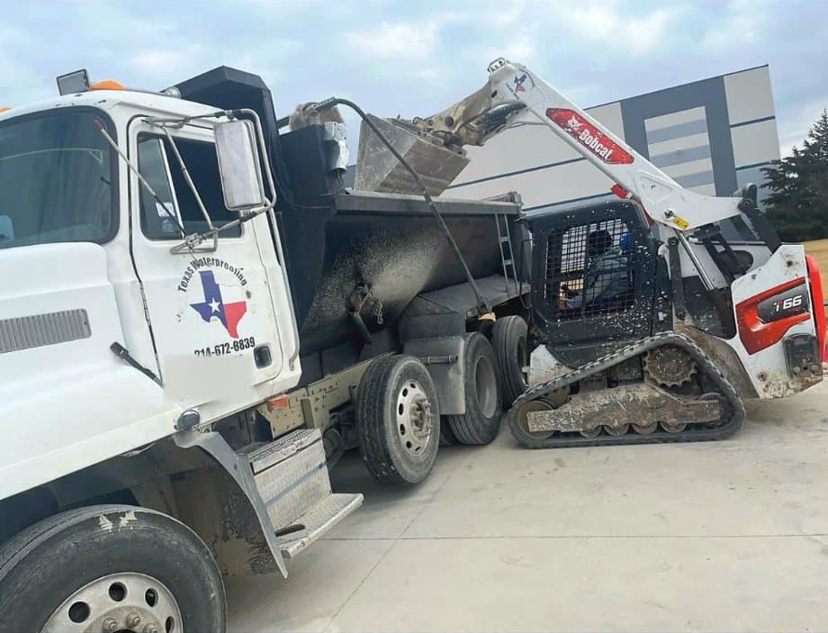 Texas Waterproofing dump truck with the company logo on the door being loaded with concrete debris by a compact track loader